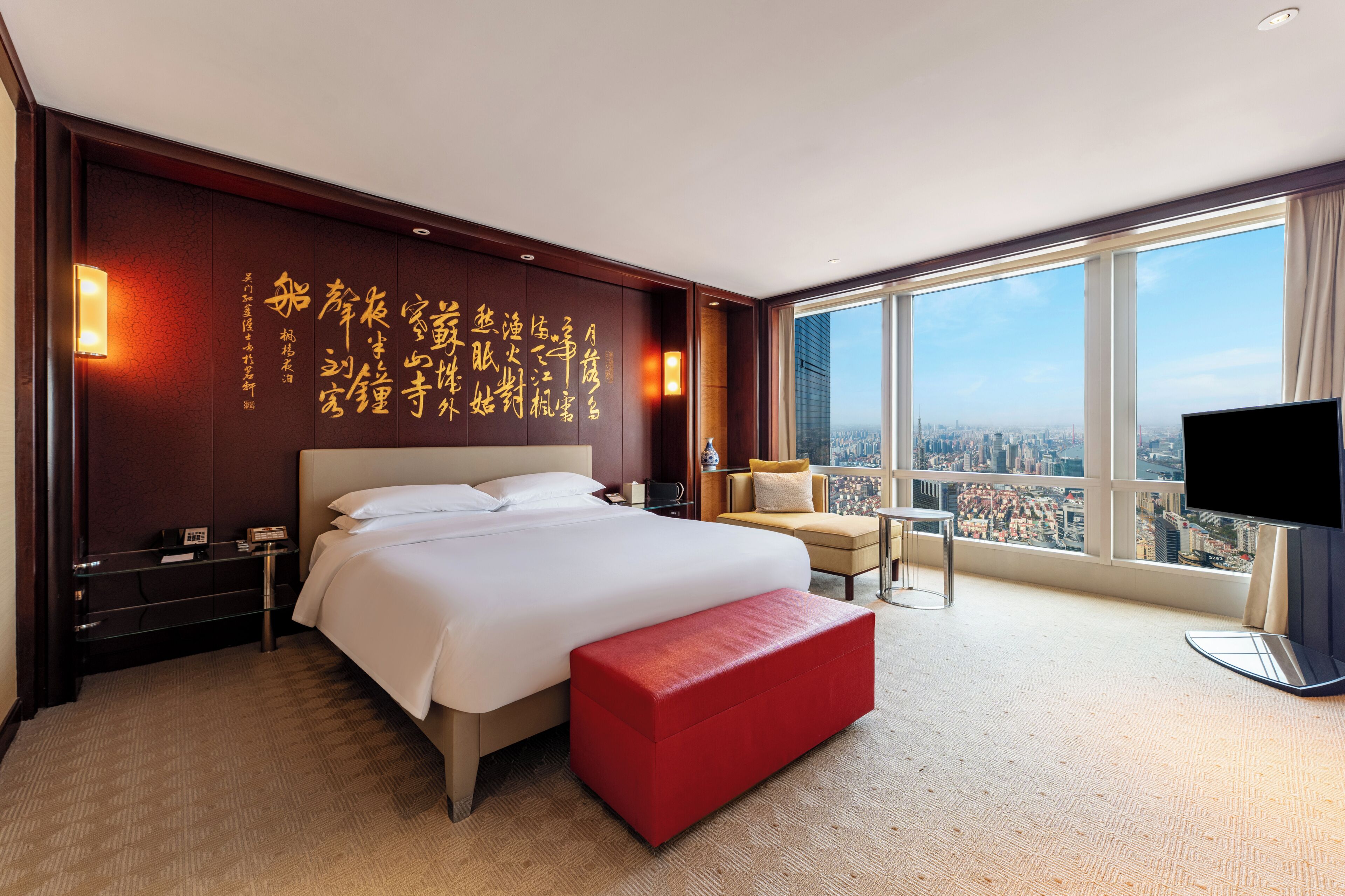 Habitación de hotel con una cama grande, un banco rojo, un televisor y vistas a la ciudad.