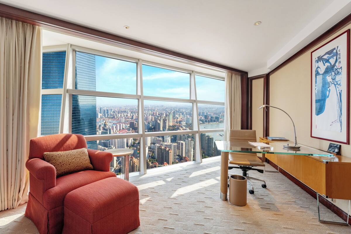 A modern hotel room with a large window, a red armchair, a glass desk, and a city view.