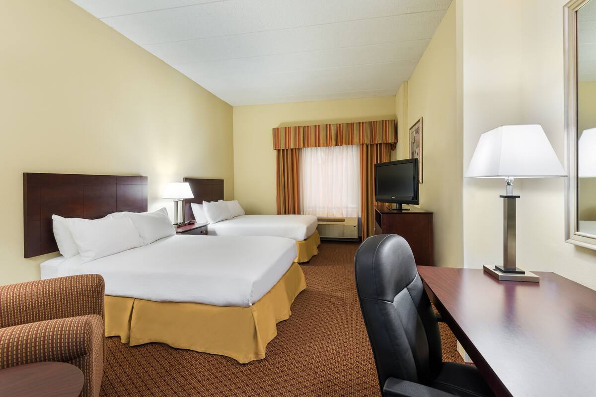 A hotel room with two beds, a desk, a chair, a TV, and a lamp.