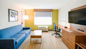 A hotel room with a blue sofa, a wooden desk, a television, a lamp, and a window with blinds. - Holiday Inn Express Richmond Airport by IHG (Sandston)