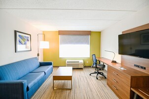 Habitación de hotel con sofá azul, escritorio de madera, televisor, lámpara y ventana con persianas.