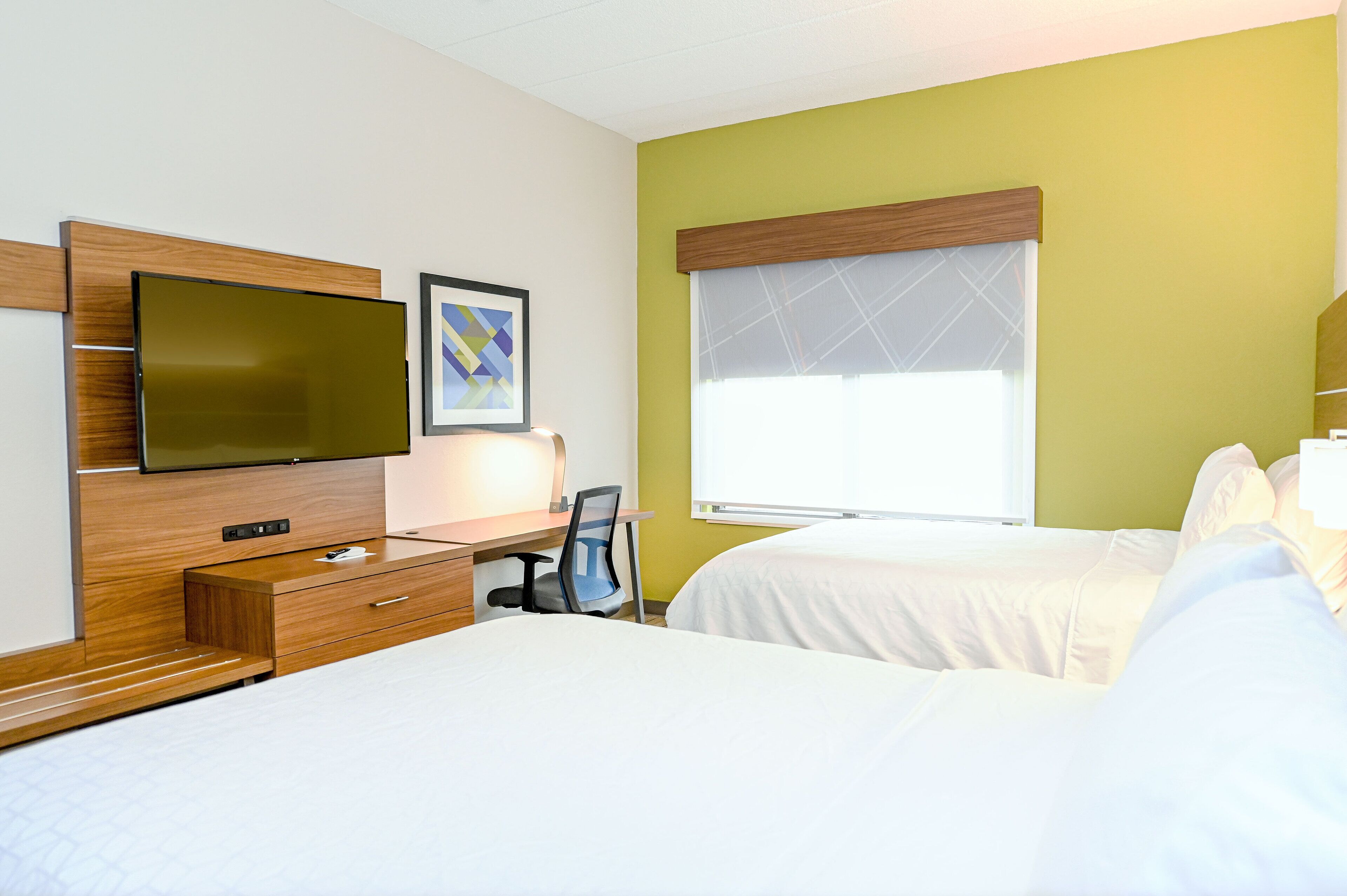 A hotel room with a bed, a desk with a chair, a television, and a window with blinds.