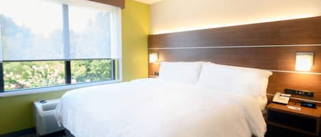 A hotel room with a large bed, a bedside table with a phone and a digital clock, a window with a view of greenery, and a wall-mounted light fixture.