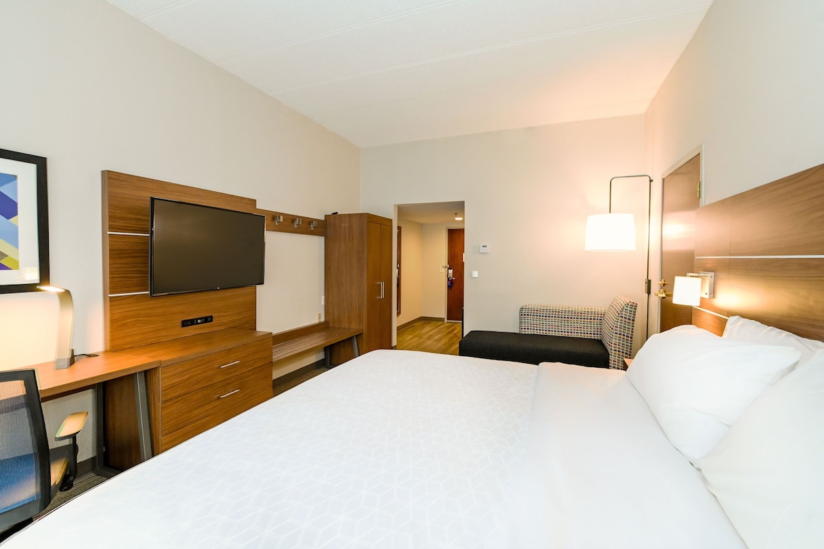 Quarto de hotel com uma cama grande, uma escrivaninha com televisão de tela plana, uma cadeira, uma mesinha de cabeceira e uma luminária.
