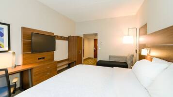 A hotel room with a large bed, a desk with a flat-screen TV, a chair, a bedside table, and a lamp.