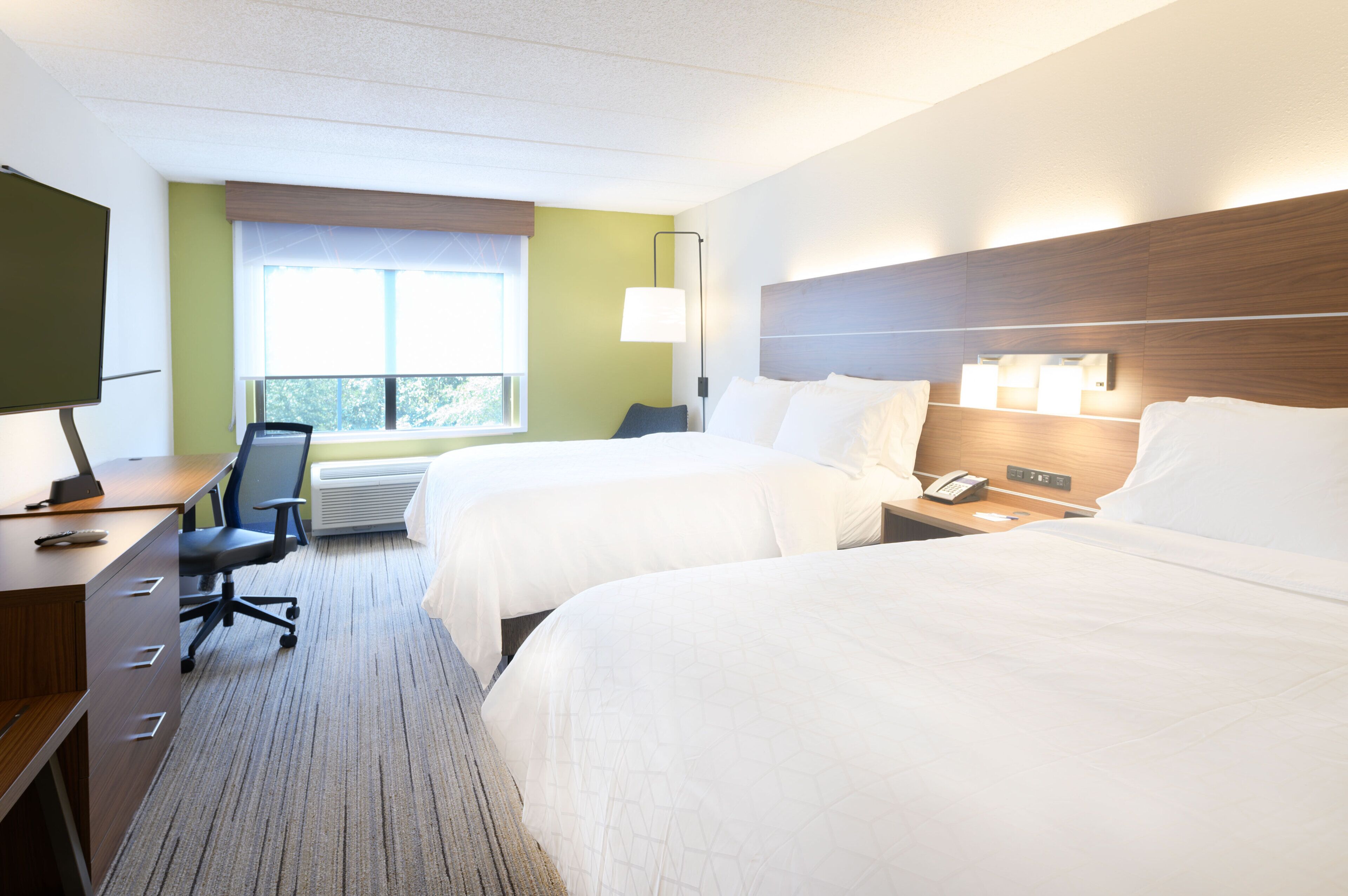 A hotel room with two beds, a desk with a chair, a TV, and a window with blinds.
