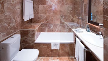 Un baño con inodoro, bañera y lavamanos, todo con paredes de mármol y elementos blancos.