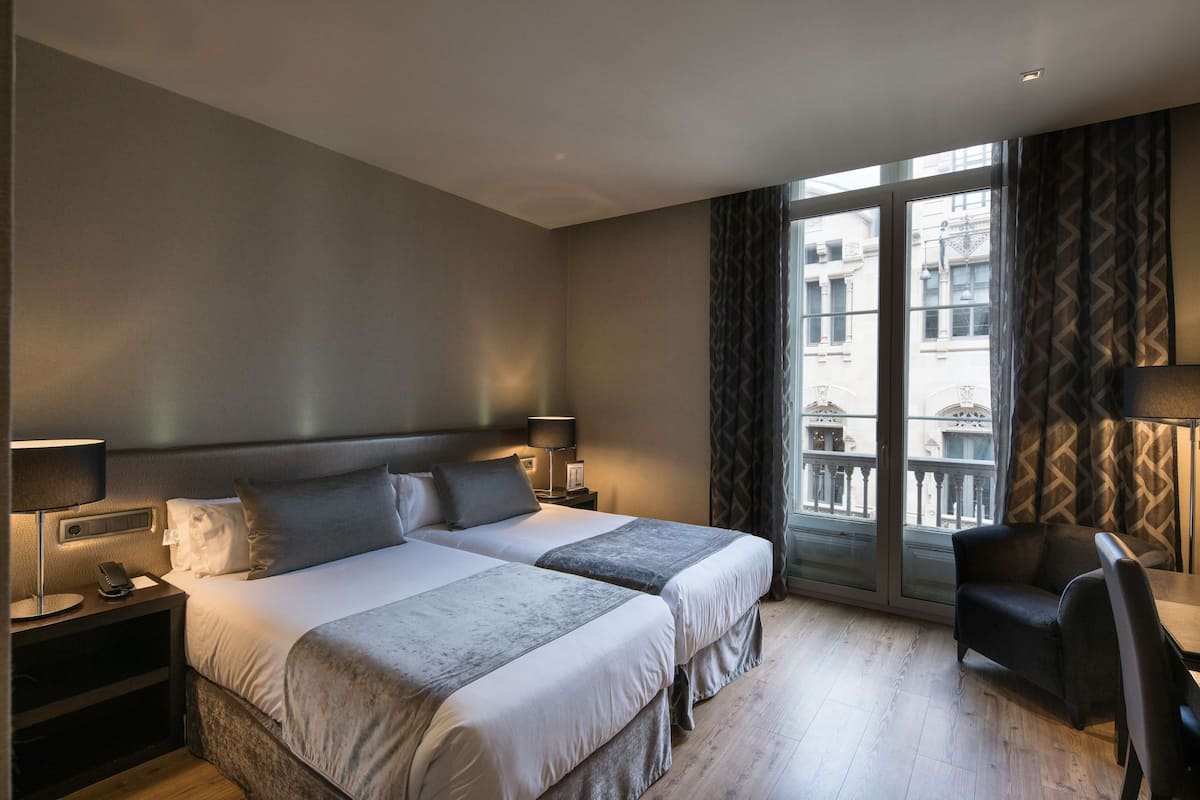 A hotel room with two beds, a chair, a desk, and a large window with a view of buildings.