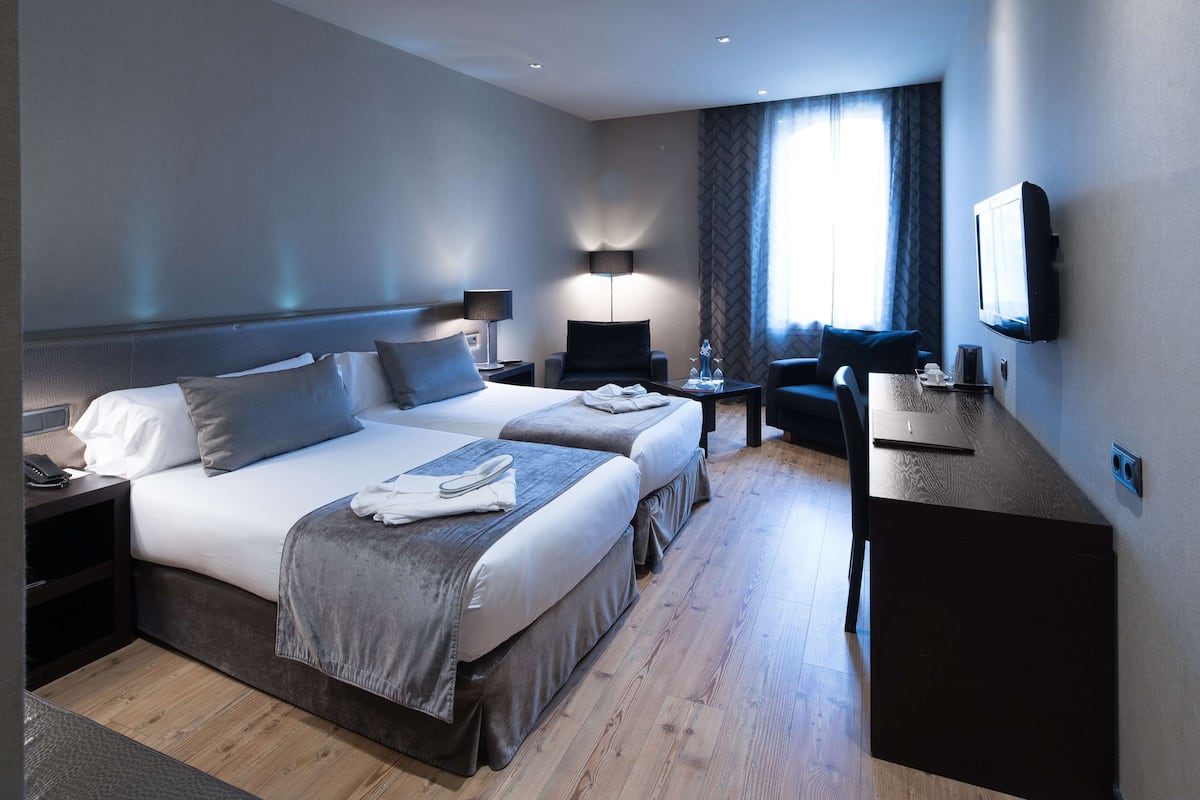 A hotel room with two beds, a desk, a chair, a TV, and a lamp.