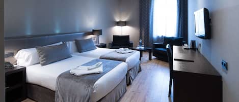 A hotel room with two beds, a desk, a chair, a TV, and a lamp.