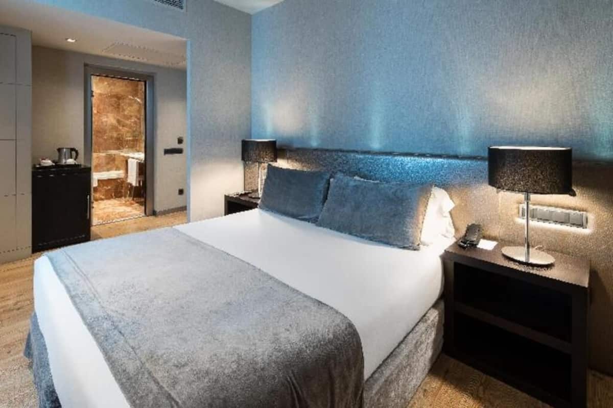 Habitación de hotel moderna con una cama grande, dos mesitas de noche con lámparas, una puerta que da al baño y vistas del pasillo.
