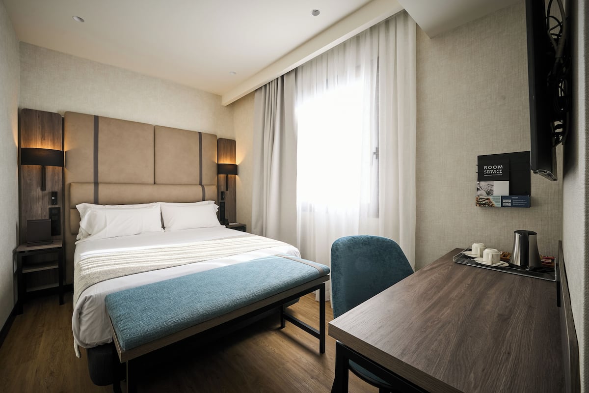 A hotel room with a bed, bedside table, lamp, chair, and a desk with a tray of items.