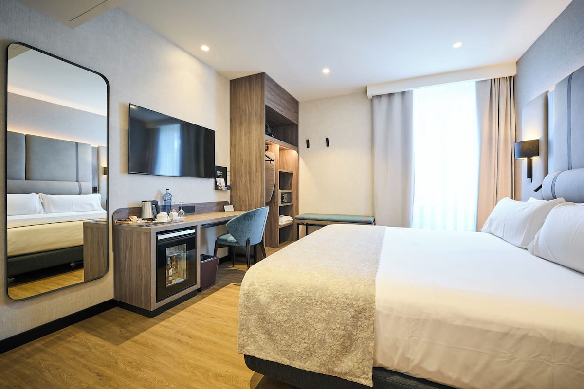 A hotel room with a bed, a desk with a chair, a TV, a mirror, and a wardrobe.