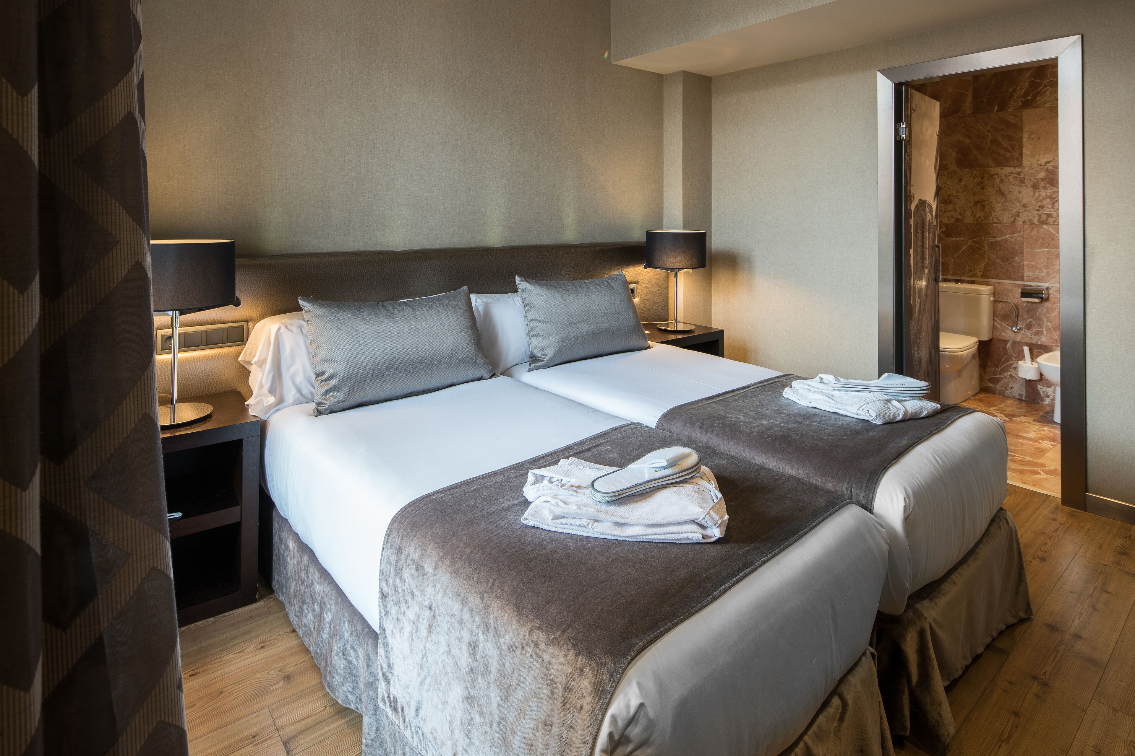 Habitación de hotel con dos camas, una mesita de noche con lámpara, un baño visible a través de una puerta abierta, y una cama con sábanas impecables y toallas.