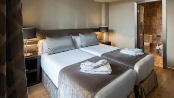 Habitación de hotel con dos camas, una mesita de noche con lámpara, un baño visible a través de una puerta abierta, y una cama con sábanas impecables y toallas.