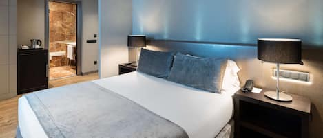 Una habitación de hotel moderna con una cama grande, dos mesitas de noche y un baño visible a través de una puerta abierta.