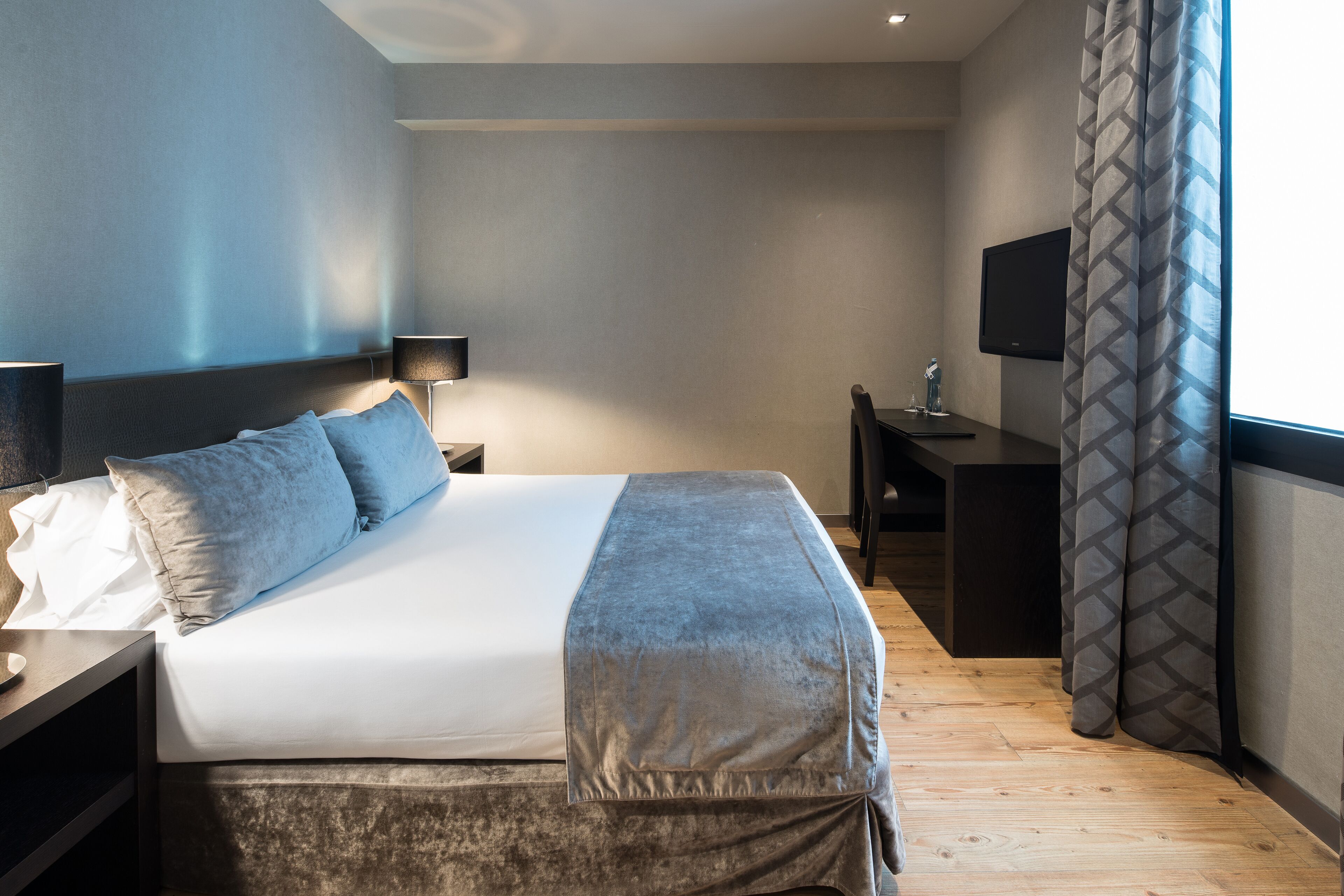 A modern hotel room with a large bed, bedside table, lamp, TV, and a desk with a chair.
