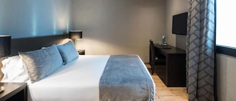 A modern hotel room with a large bed, bedside table, lamp, TV, and a desk with a chair.