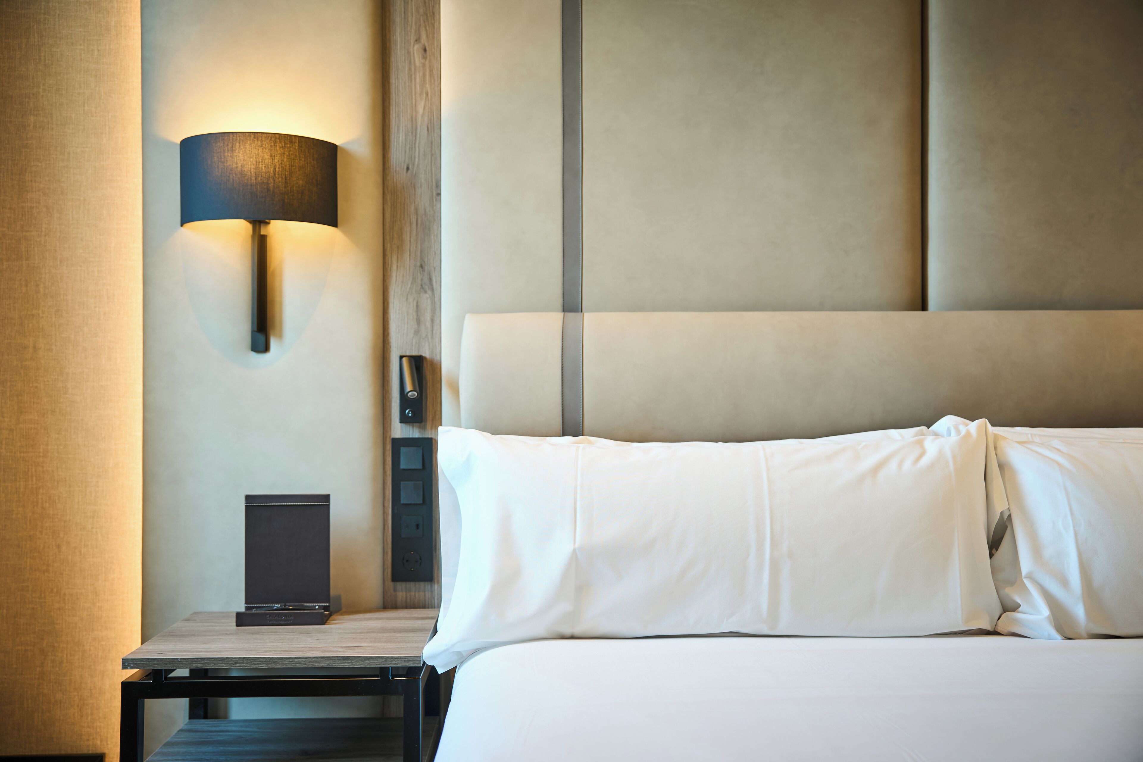 A hotel room with a bed, a nightstand, a lamp, and a control panel.