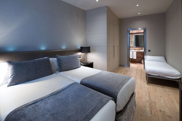 A modern hotel room with two beds, a nightstand, a lamp, and a bathroom visible through an open door.