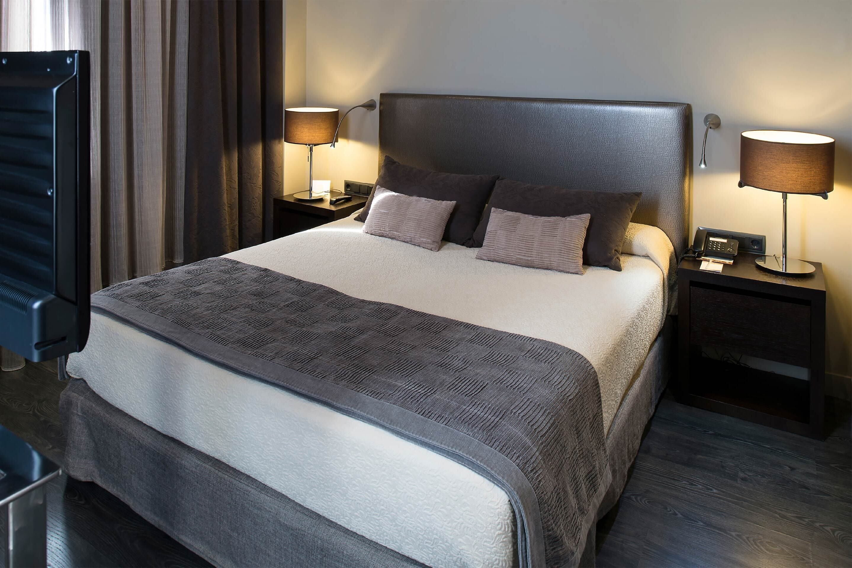 A neatly made bed with a grey and white duvet, two bedside lamps, and a nightstand with a phone and a book.