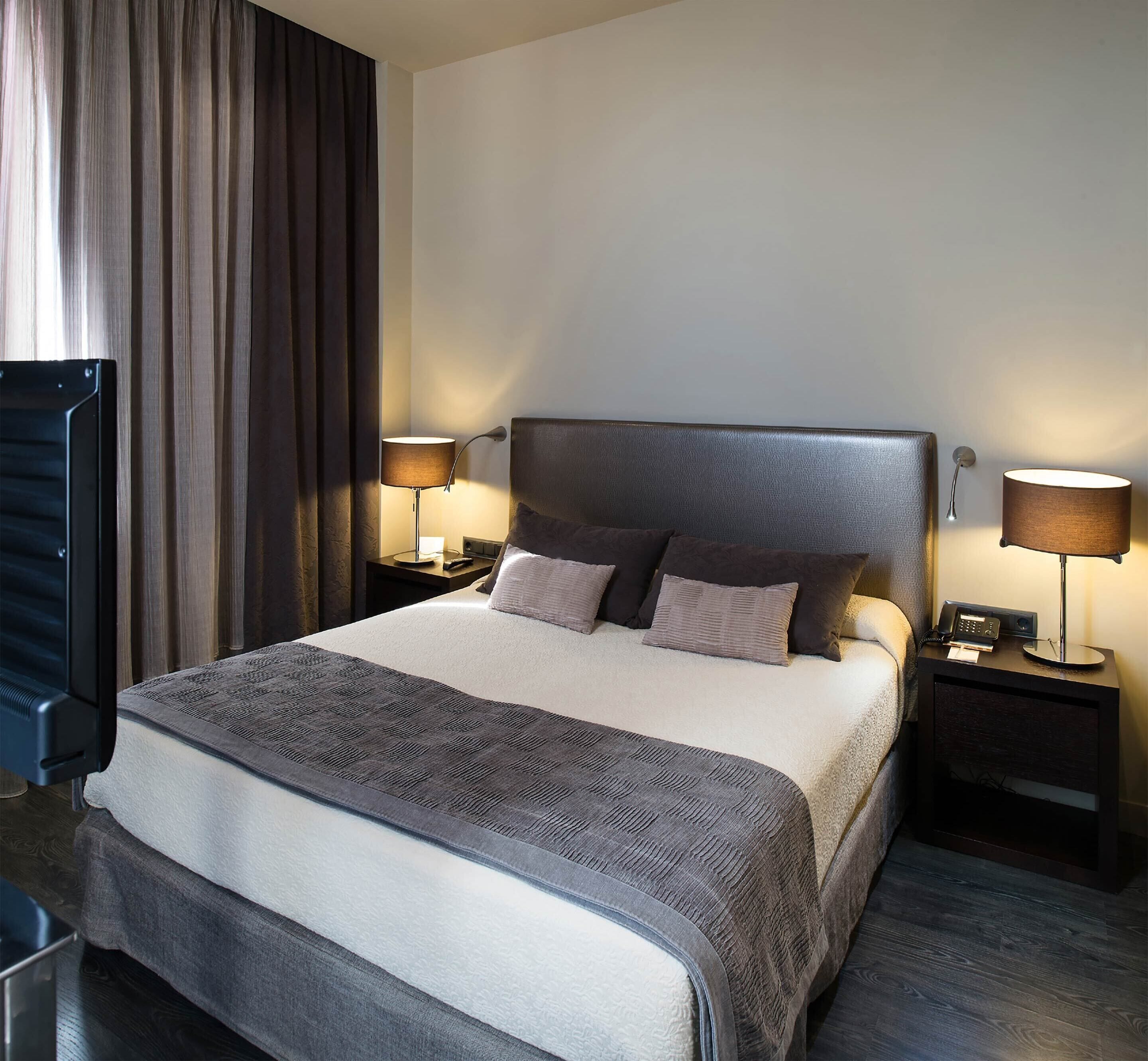 A hotel room with a large bed, two bedside tables with lamps, a dark headboard, and a view of a curtain and a wall.