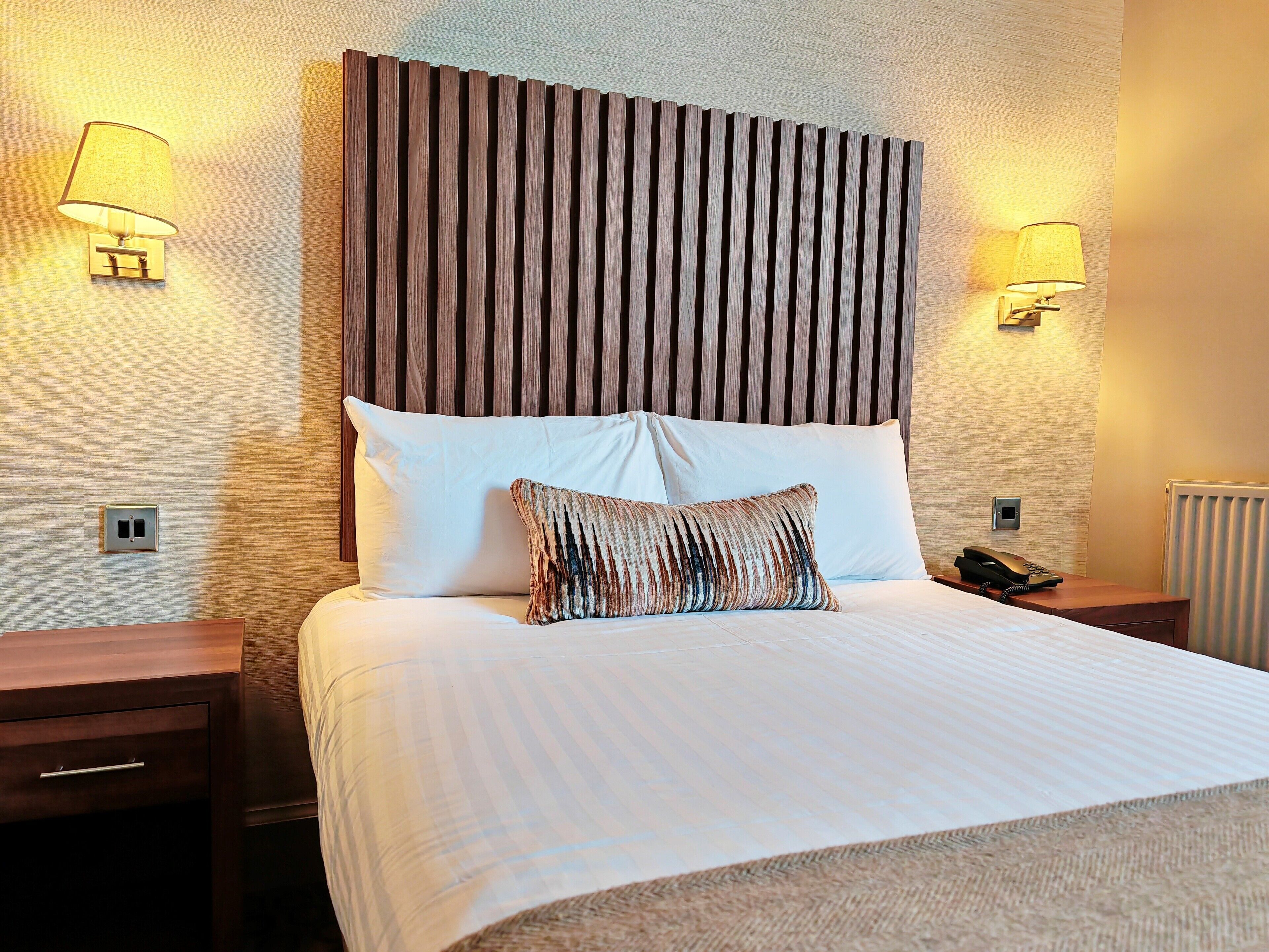 Une chambre d’hôtel avec un grand lit, deux lampes de chevet, une tête de lit en bois et un téléphone sur la table de chevet.