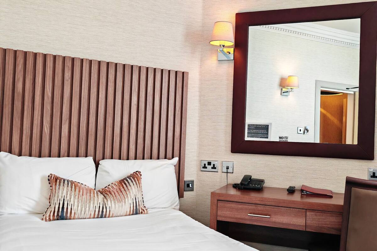 Una habitación de hotel con una cama grande, un cabecero de madera, una mesita de noche con una lámpara y un espejo en la pared.
