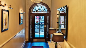 Un couloir d’hôtel avec un lustre, un tapis rouge d’accueil, un miroir et une porte donnant sur la rue.