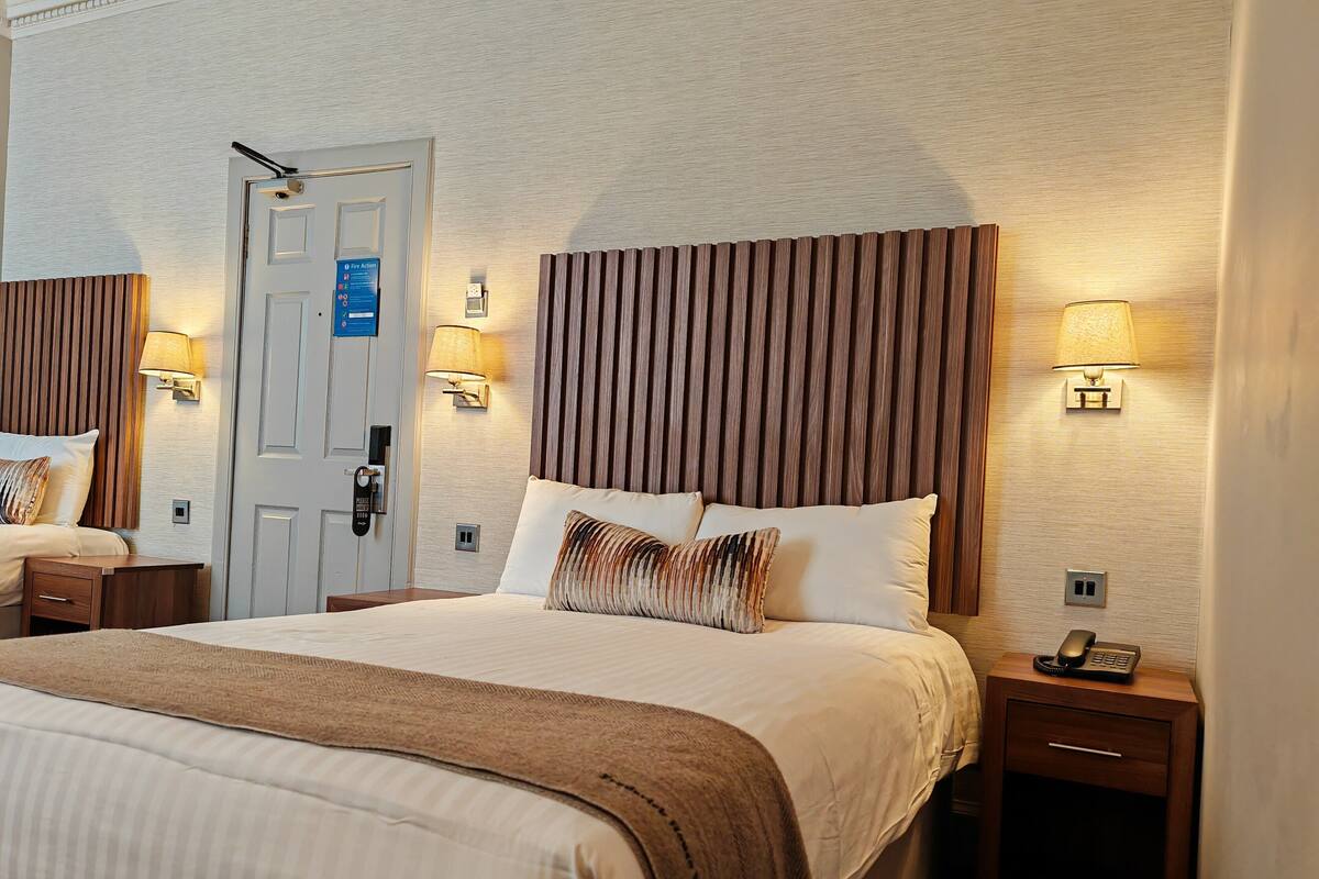 Una habitación de hotel con una cama grande, dos mesitas de noche, una puerta con lector de tarjeta y lámparas de pared.