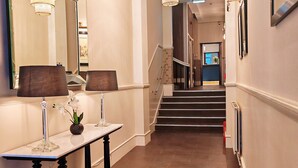 Un couloir avec une console en marbre, deux lampes, un vase de fleurs, un lustre et un escalier menant à un autre espace.