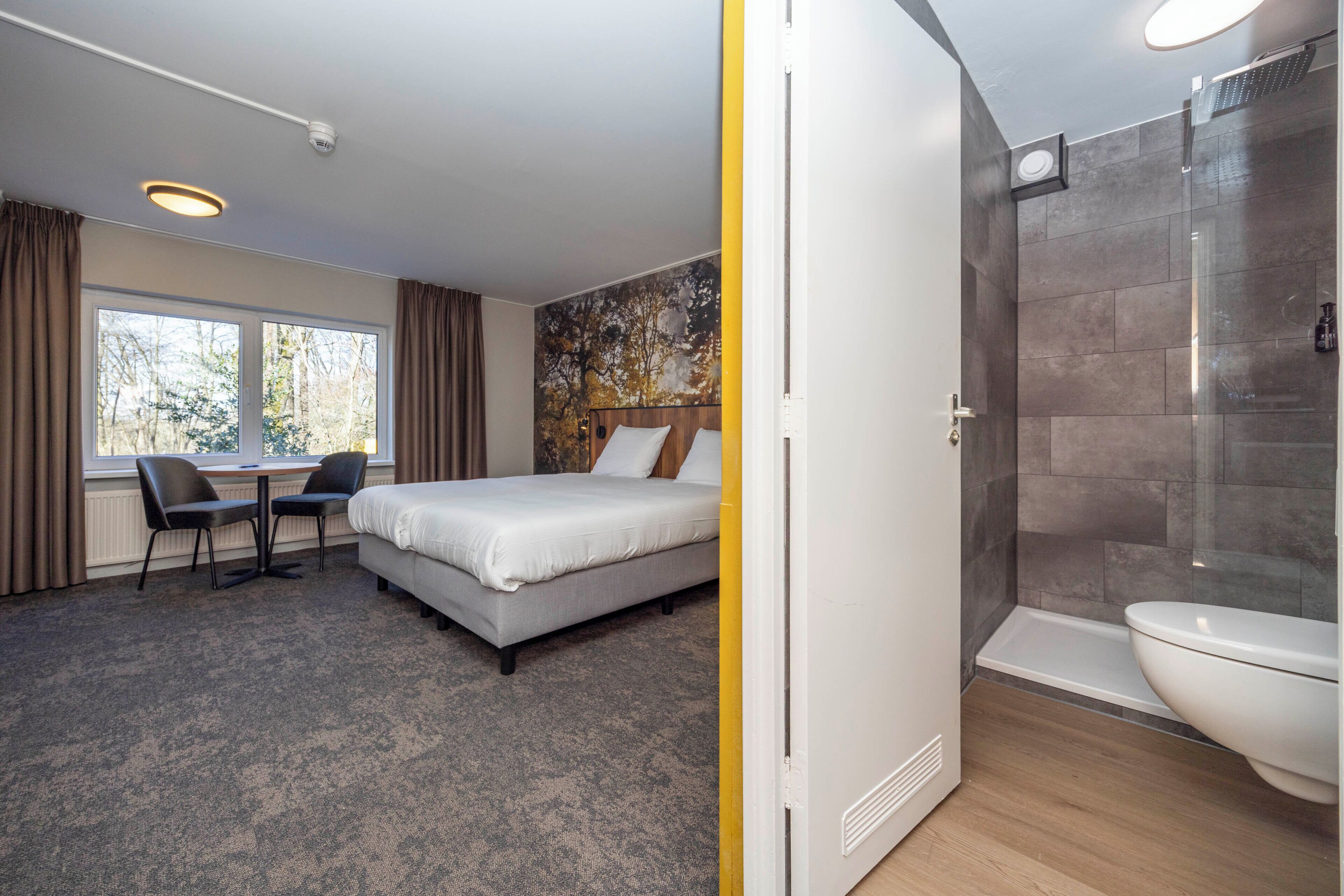 A hotel room with a bed, a desk with chairs, a window with curtains, a shower area, and a toilet.