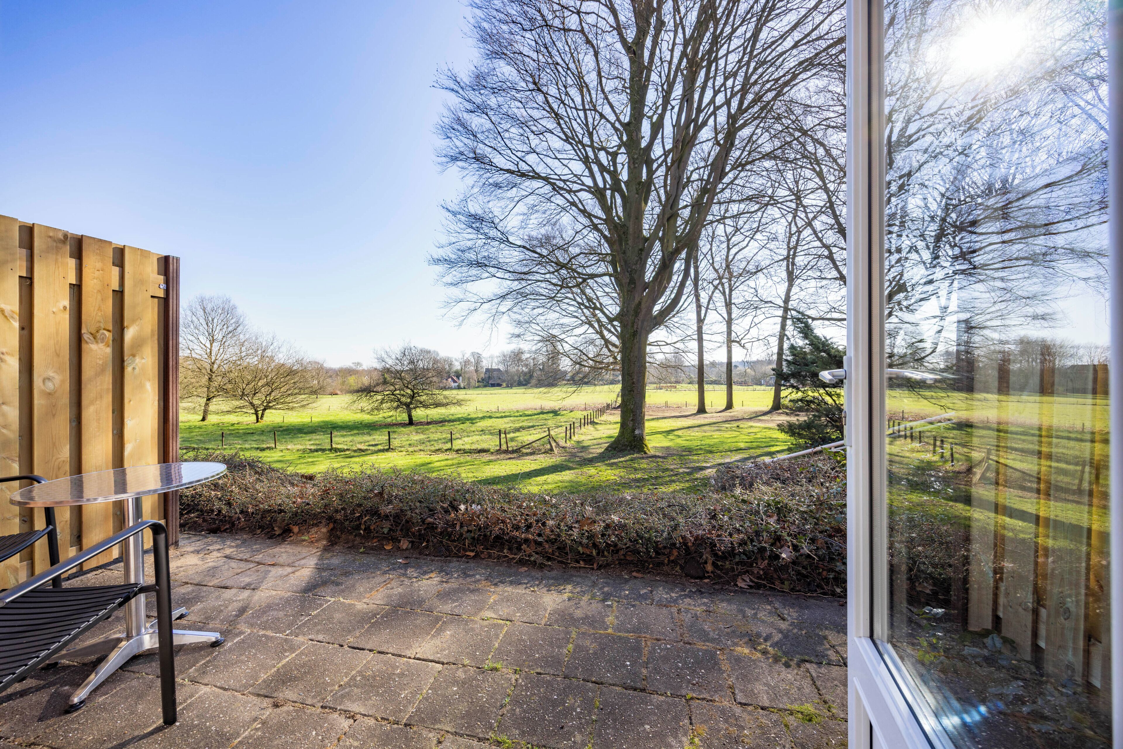 Eine Terrasse mit Tisch und Stühlen, Blick auf ein grünes Feld und einen großen Baum.