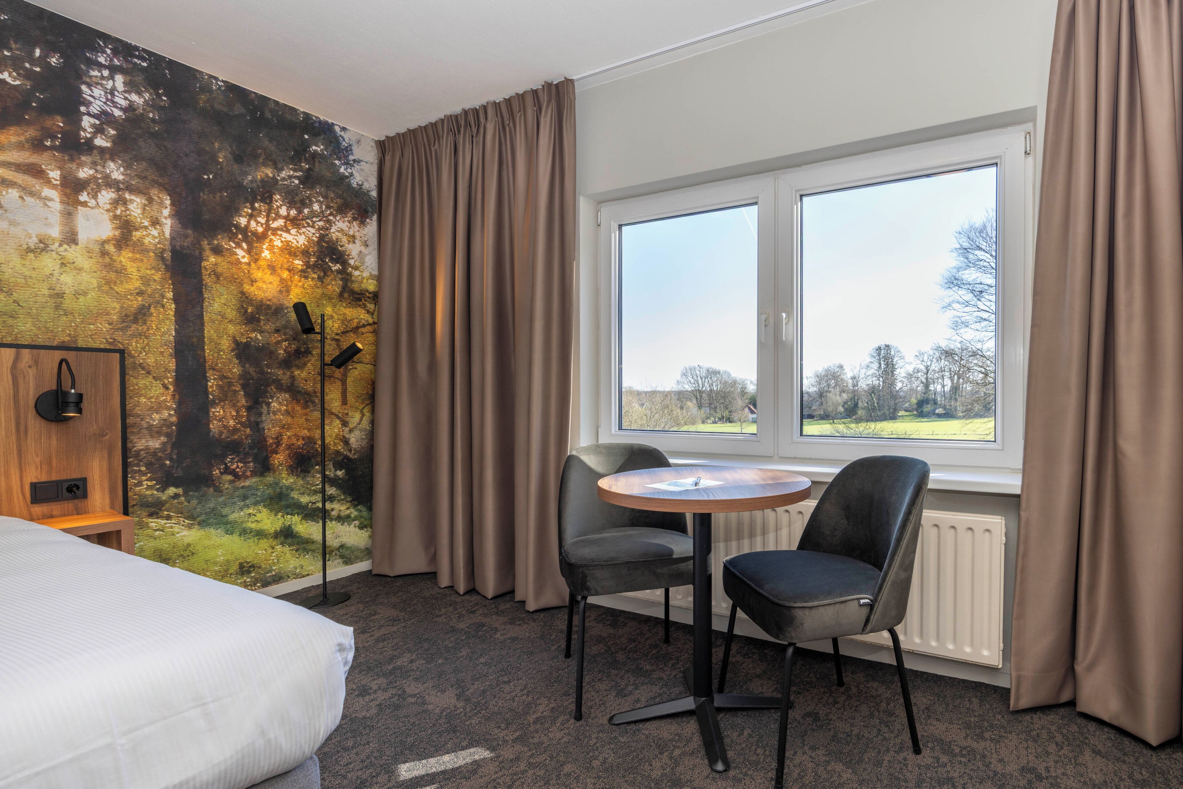 A hotel room with a bed, a round table with two chairs, a radiator, and a large window with curtains.