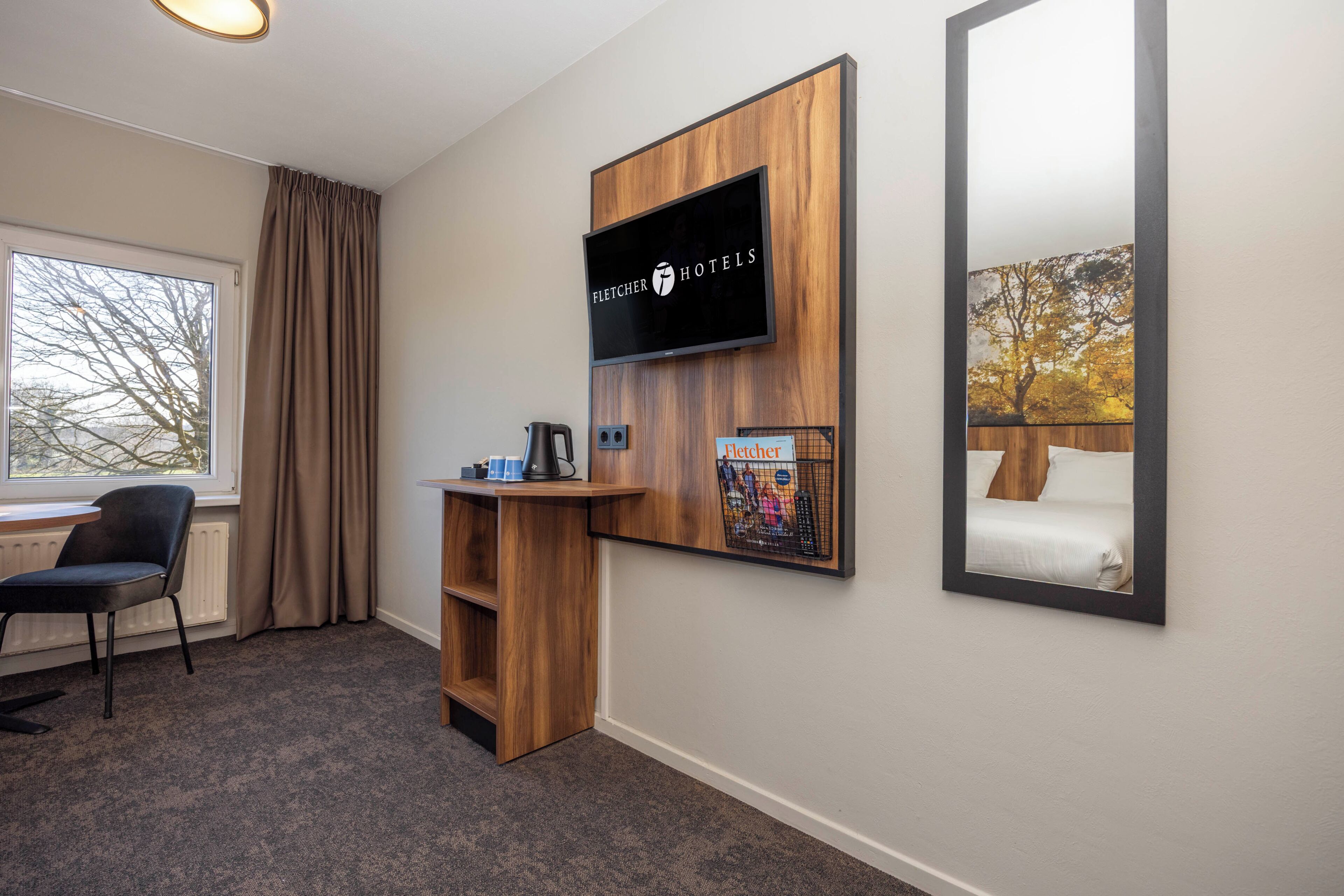 A hotel room with a large window, a desk with a kettle, a chair, a mounted television, and a framed picture on the wall.