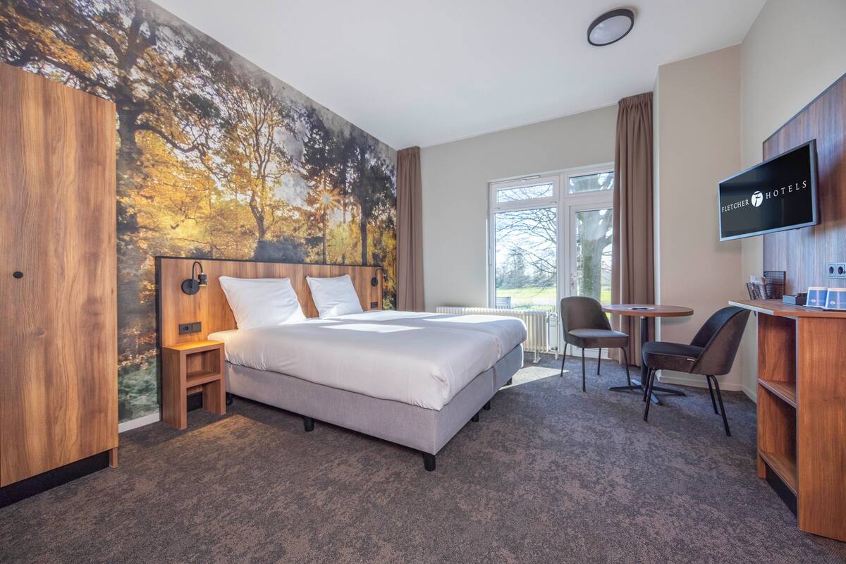 Une chambre d’hôtel avec un grand lit, un bureau, une chaise, une télévision et une fresque représentant des arbres.