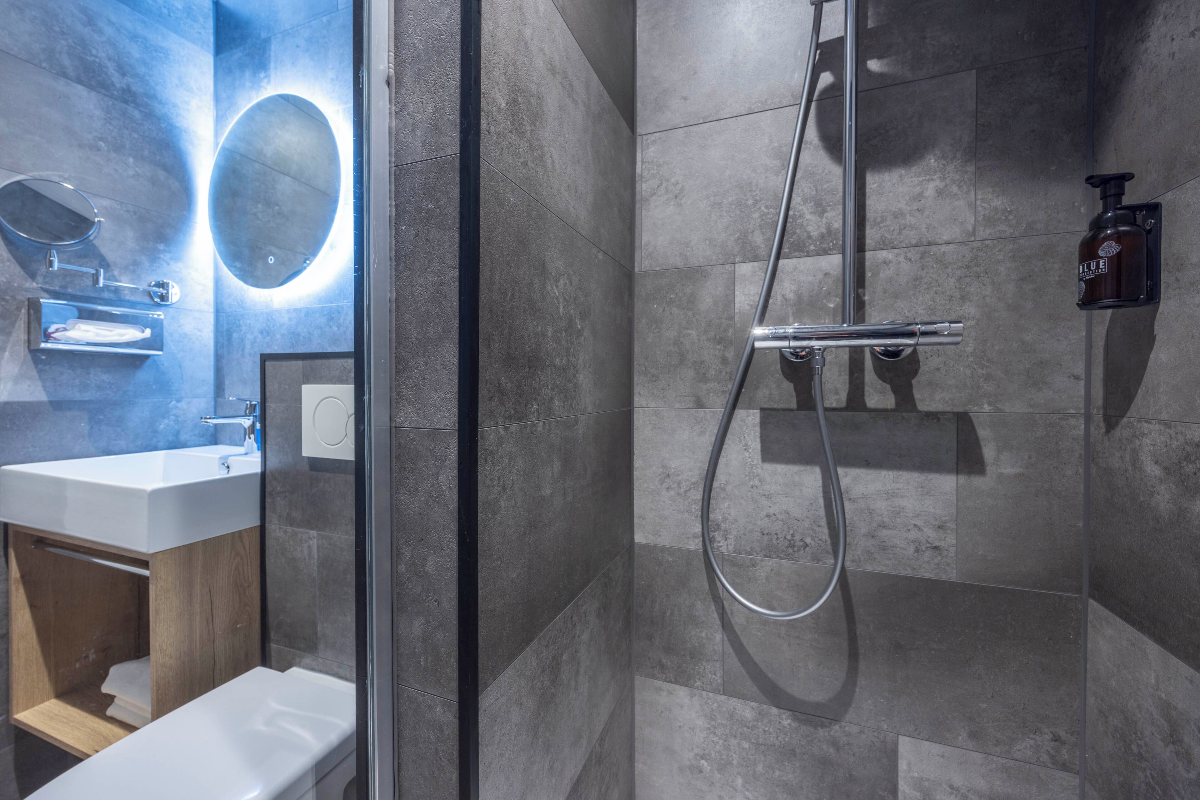 Un baño moderno con ducha de vidrio, lavamanos blanco y espejo con luz circular.