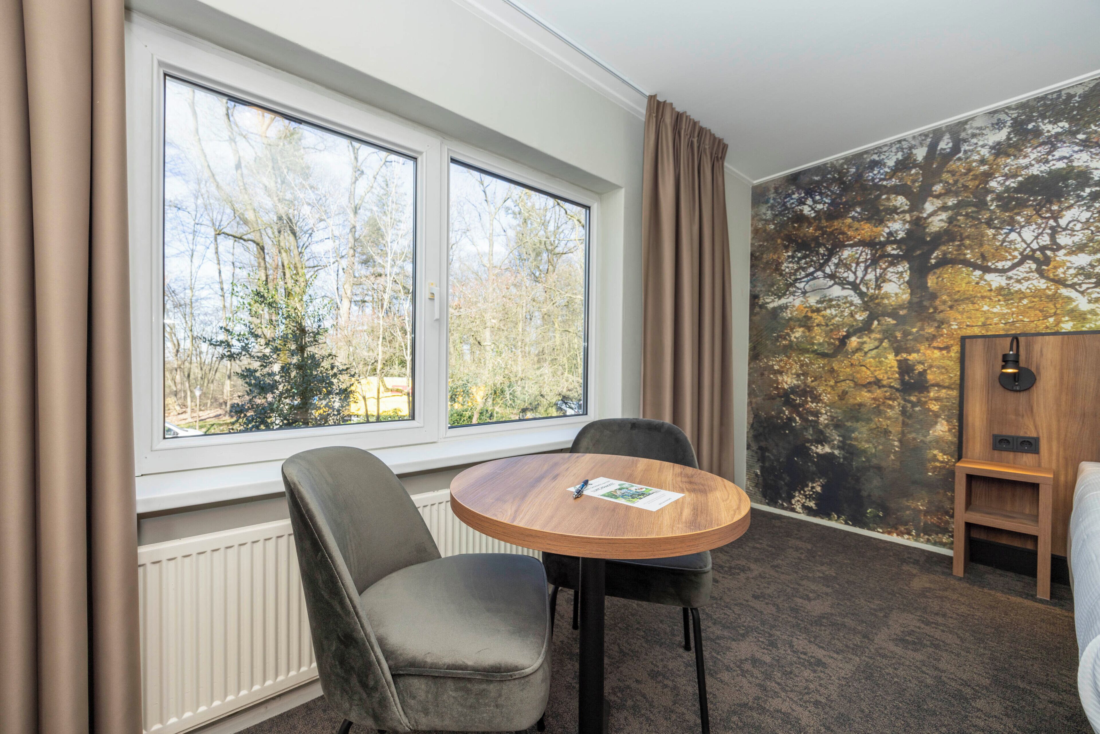 Une pièce avec une table ronde en bois, deux chaises grises, un radiateur, une fenêtre avec des rideaux et une fresque murale représentant des arbres.