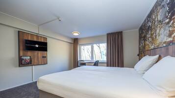 A hotel room with a bed, a desk, a chair, a window with curtains, and a wall-mounted television.