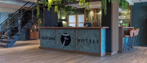 A hotel reception area with a staircase, a reception desk with a logo, and a bar area.
