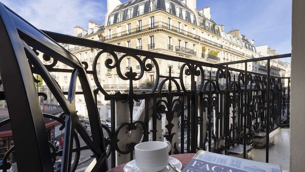 Un balcon orné de balustrades en fer forgé, une table avec une tasse et une soucoupe blanches, et un livre intitulé « FACADES PARISIENNES ».