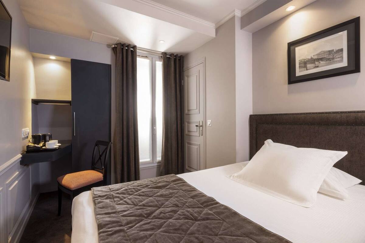 A hotel room with a bed, a desk with a chair, a framed picture on the wall, and a window with curtains.