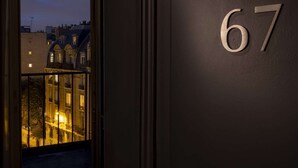 Un intérieur sombre, avec une fenêtre donnant sur un bâtiment illuminé à l’extérieur. Le numéro 67 est affiché sur le mur.