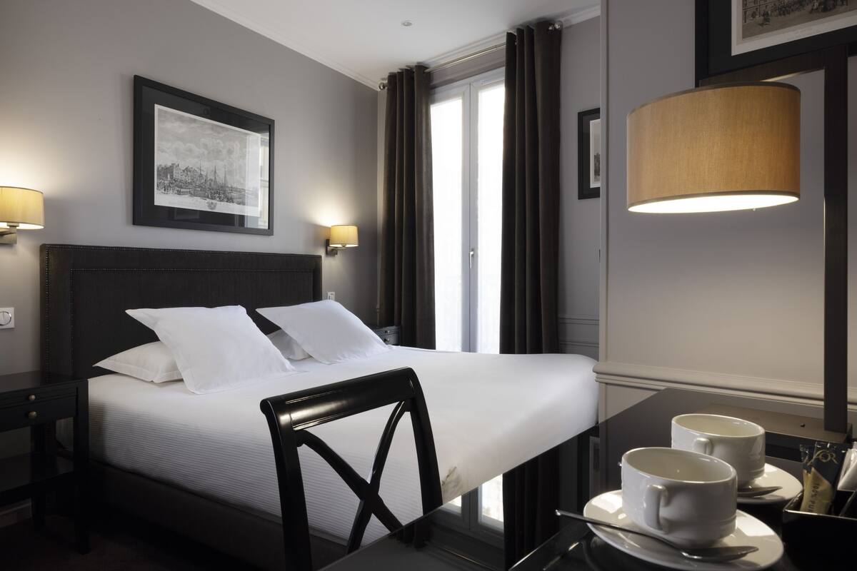 A hotel room with a large bed, two bedside lamps, a framed picture on the wall, a window with curtains, and a table with two cups and a spoon.