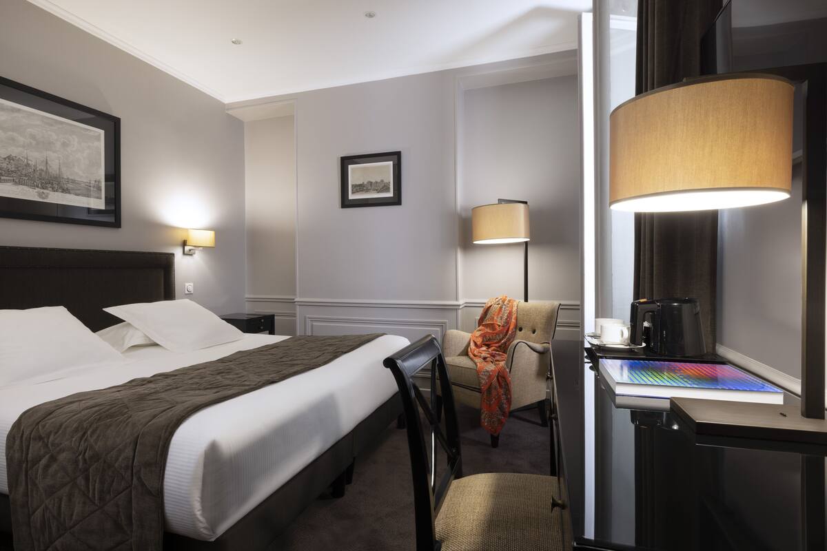 A hotel room with a bed, a desk with a lamp, a chair, and a coffee maker.