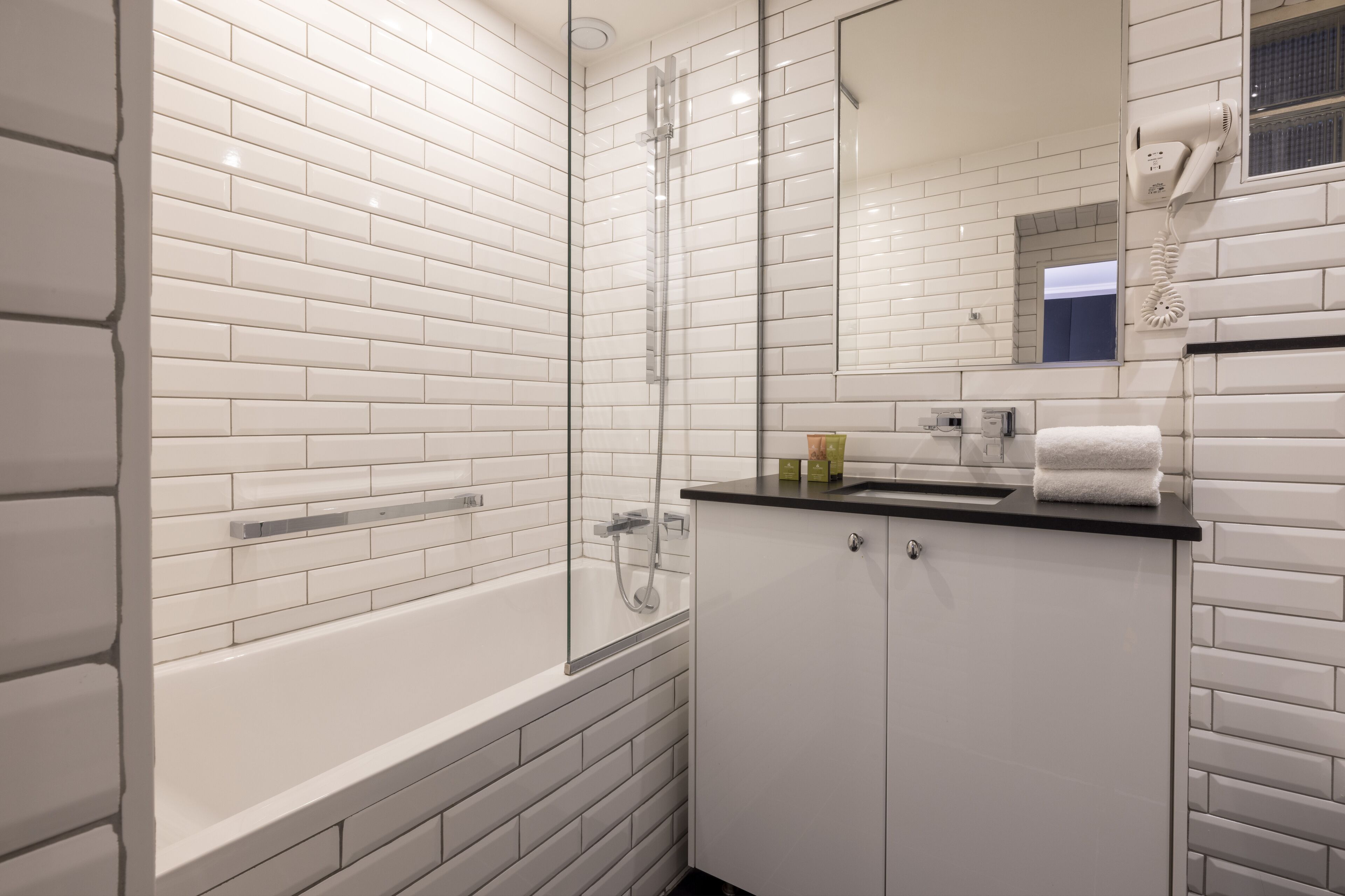 Une salle de bain moderne avec des carreaux de céramique blancs rectangulaires, une douche en verre, un comptoir noir et un miroir avec un sèche-cheveux.