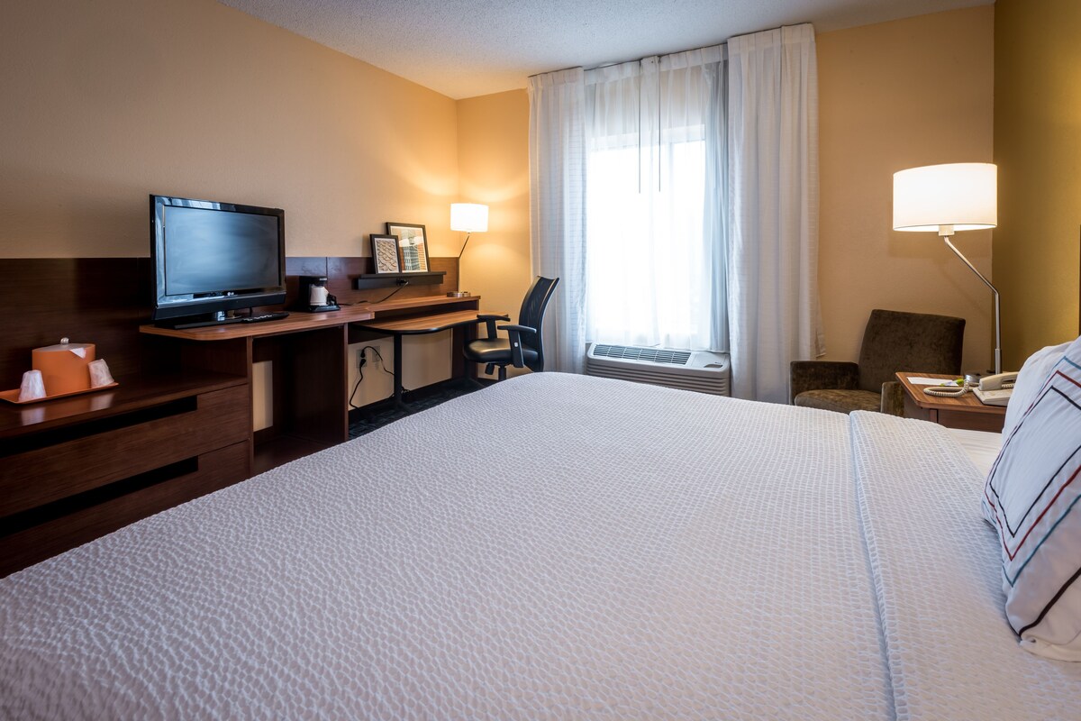 A hotel room with a large bed, a desk with a television, a chair, a lamp, and a window with curtains.