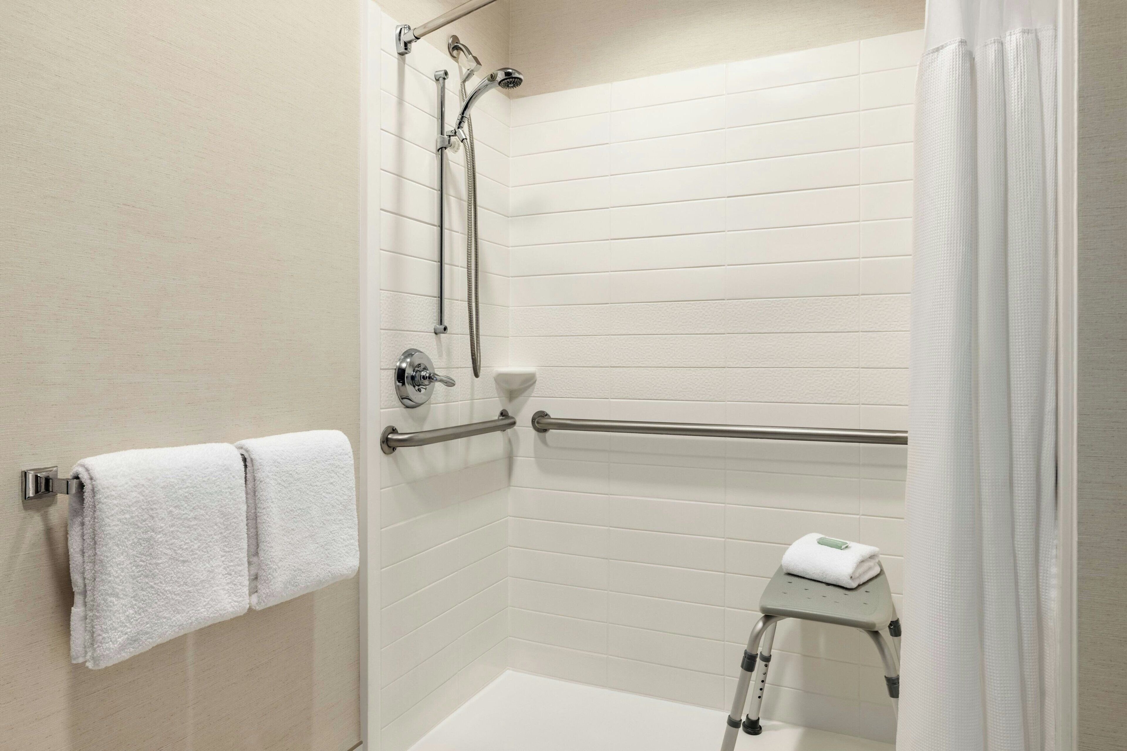 A modern bathroom with white tiled walls, a shower with a handheld showerhead, a grab bar, and a small step stool with a towel.