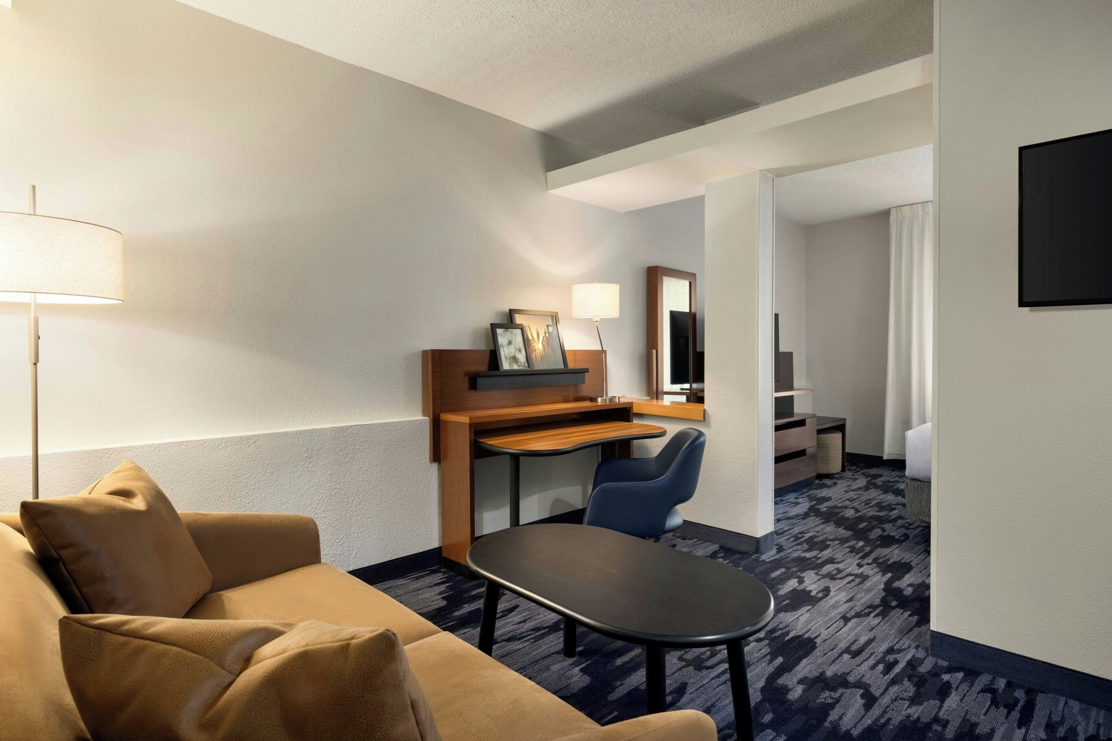 A hotel room with a sofa, a small table, a desk with a chair, a lamp, a mirror, and a TV.