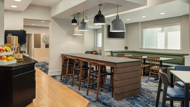 A modern hotel lobby with a central bar area, seating arrangements, and a large window providing natural light.